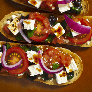 Prepara una deliciosa Bruschetta griega con queso feta, tomates, aceitunas griegas deshuesadas y orégano sobre rebanadas crujientes de pan casero. ¡Un auténtico e irresistible aperitivo mediterráneo!