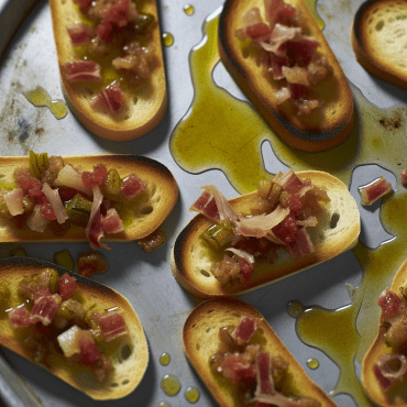 Receta sarda: Crostinis de pan carasau con queso rasco y miel. Un tentempié sardo auténtico y rápido donde el queso rasco se funde delicadamente sobre el crujiente pan carasau, realzado por la dulzura de la miel de castaño. Una combinación perfecta de sabores para deleitar a sus invitados.